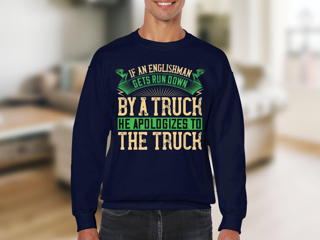 A person wears a Garment Graphics navy blue sweatshirt with bold, humorous text: If an Englishman gets run down by a truck, he apologizes to the truck. The green and beige lettering and decorative elements pop on this unisex piece against a blurred indoor backdrop.