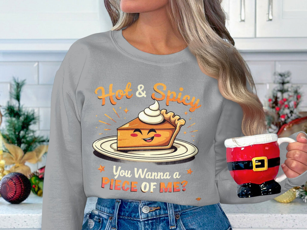 A person with long hair holds a Santa mug, wearing a festive Garment Graphics unisex gray pie graphic sweatshirt that reads Hot & Spicy, You Wanna a Piece of Me? Perfectly suited for holiday decor with ornaments and greenery.