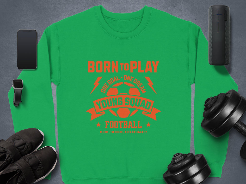 The unisex green football sweatshirt by Garment Graphics features red text: Born to Play, One Goal - One Dream, Young Squad Football, Kick. Score. Celebrate! It is surrounded by a smartphone, smartwatch, portable speaker, dumbbells, and black shoes on a gray background—ideal for young squads!.