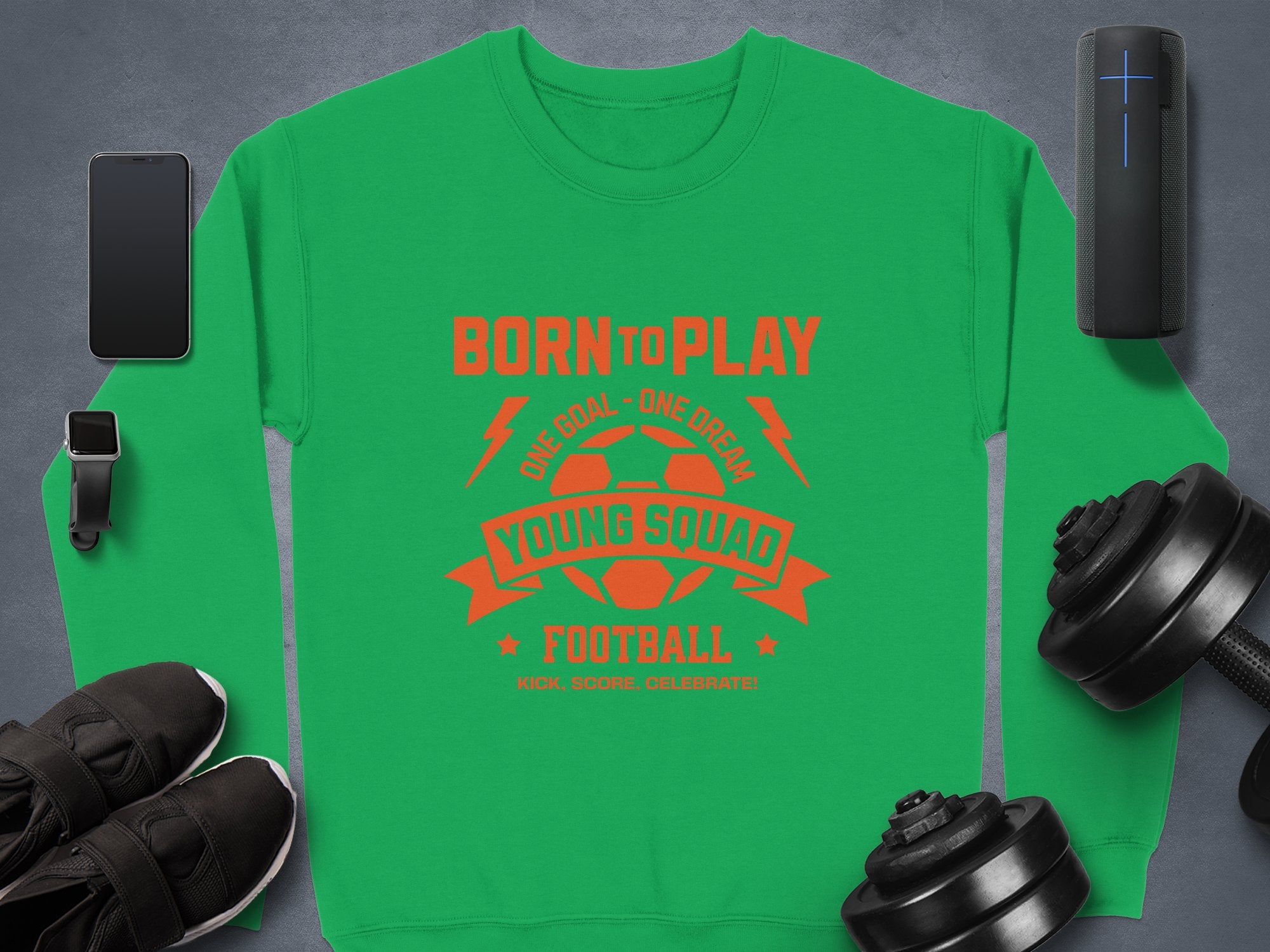 The unisex green football sweatshirt by Garment Graphics features red text: Born to Play, One Goal - One Dream, Young Squad Football, Kick. Score. Celebrate! It is surrounded by a smartphone, smartwatch, portable speaker, dumbbells, and black shoes on a gray background—ideal for young squads!.