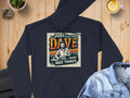 A black medium-heavy hoodie featuring Just Dave Doing Dave Things from Garment Graphics sits on a wooden table beside a denim jacket, latte, small plant, and phone.