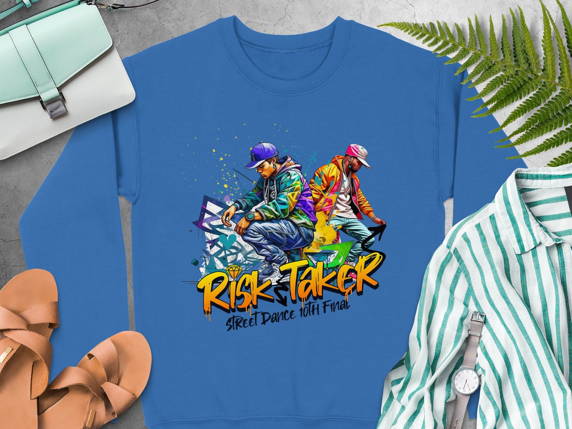 A blue sweatshirt by Garment Graphics features two dancers in lively streetwear striking bold poses with Risk Taker Street Dance 10th Final printed below. Styled alongside a striped shirt, purse, shoes, and ferns, it captures the essence of street dance finals chic.