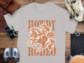 Garment Graphics offers a gray Rodeo T-shirt featuring a cowgirl on a galloping horse with Howdy Rodeo and Not a Beginner. Perfect for enthusiasts, it sits on a wooden surface surrounded by a camera, binoculars, gloves, and leather accessories.