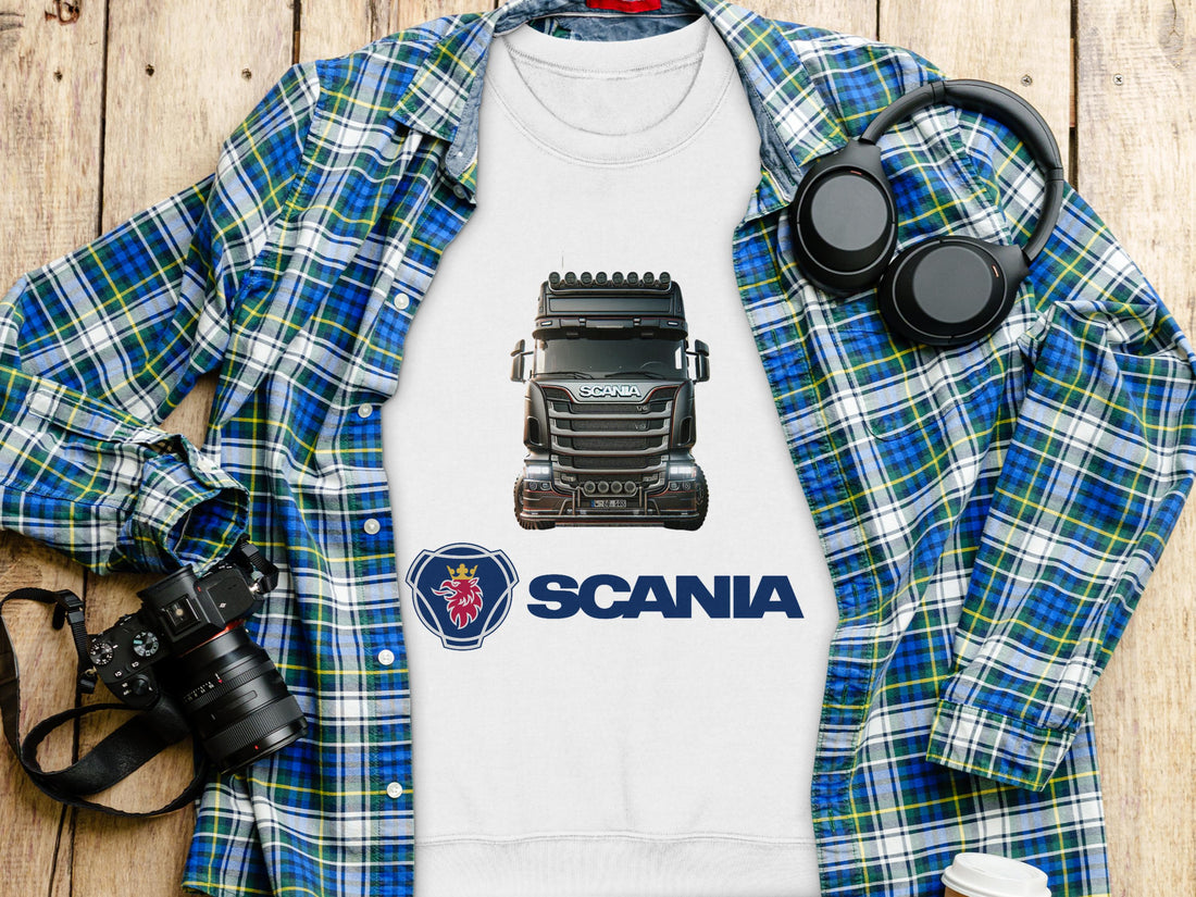 A Garment Graphics white T-shirt with a Scania truck design and logo lies on wood, alongside a blue plaid shirt, trendy sweatshirts, a camera, headphones, and a laptop—creating a casual and tech-savvy scene with graphic flair.