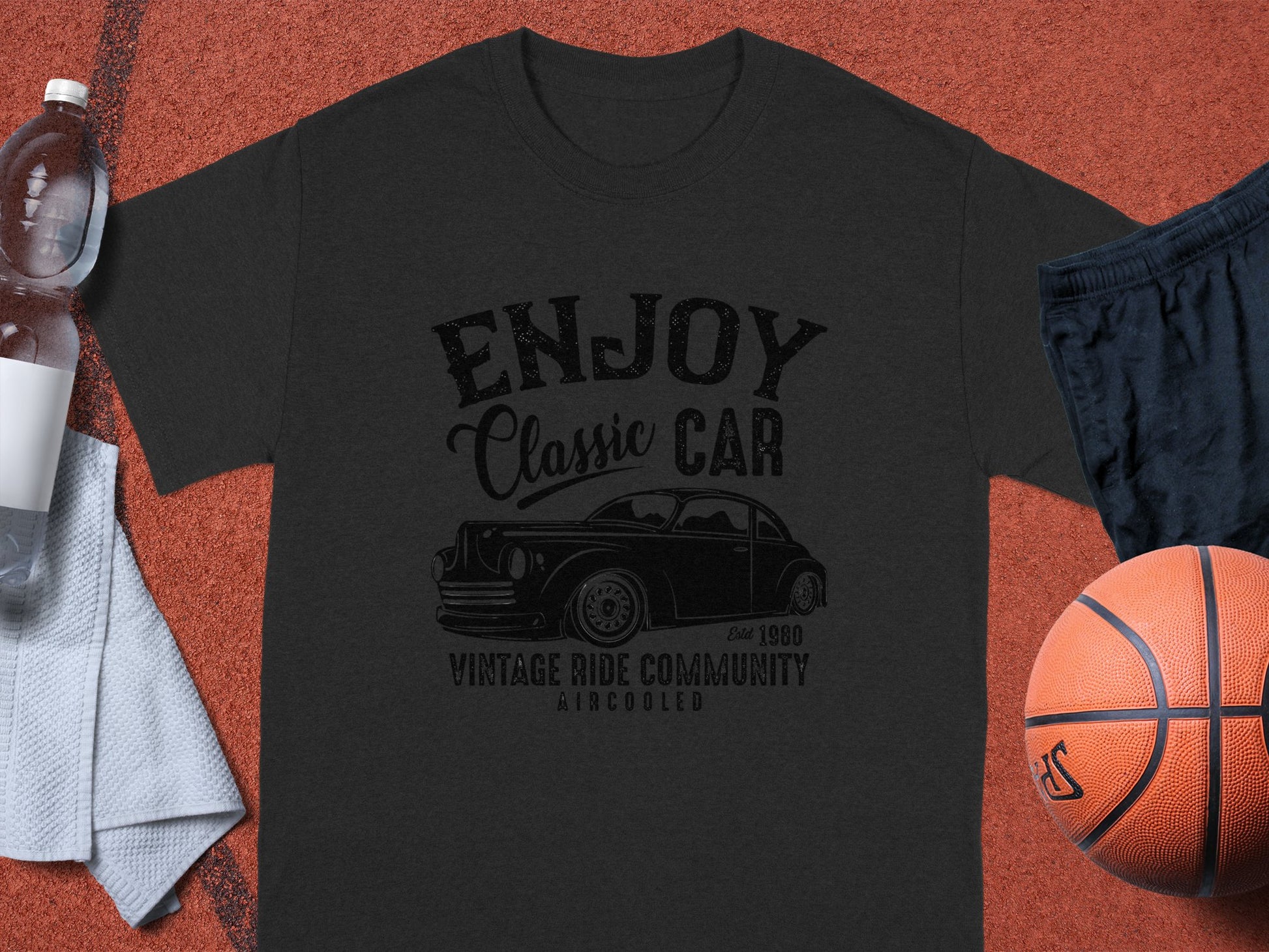 The Garment Graphics black Enjoy Classic Car T-shirt lies on an orange surface, accompanied by car enthusiast essentials: a water bottle, navy shorts, a basketball, and a white towel.