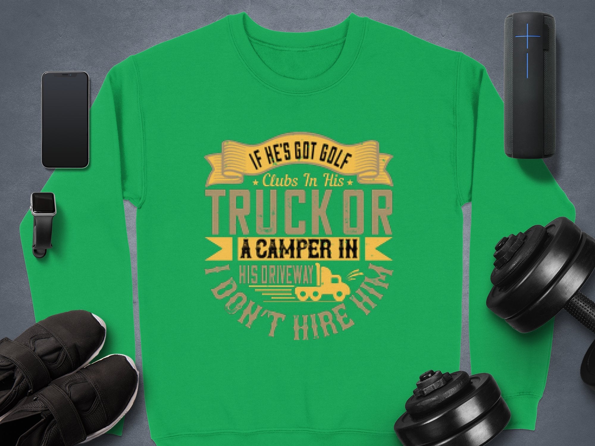 Garment Graphics presents a green sweatshirt featuring a humorous graphic: If hes got golf clubs in his truck or a camper in his driveway, I dont hire him, surrounded by a smartphone, speakers, sneakers, and dumbbells on a gray surface.
