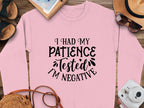 A pink Garment Graphics sweatshirt, perfect for outings, is displayed on a wooden surface. It humorously states, I had my patience tested, Im negative. Nearby are a notebook, straw hat, sunglasses, and sandals. Its unisex sizing offers comfort and style for all.