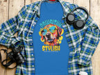 A flat lay image showcases a Garment Graphics Paw-sitive Stylish blue dog t-shirt with a vibrant canine print, alongside an open plaid shirt, headphones, a camera, and a white tumbler on rustic wood. Ideal for chilling like a canine villain.
