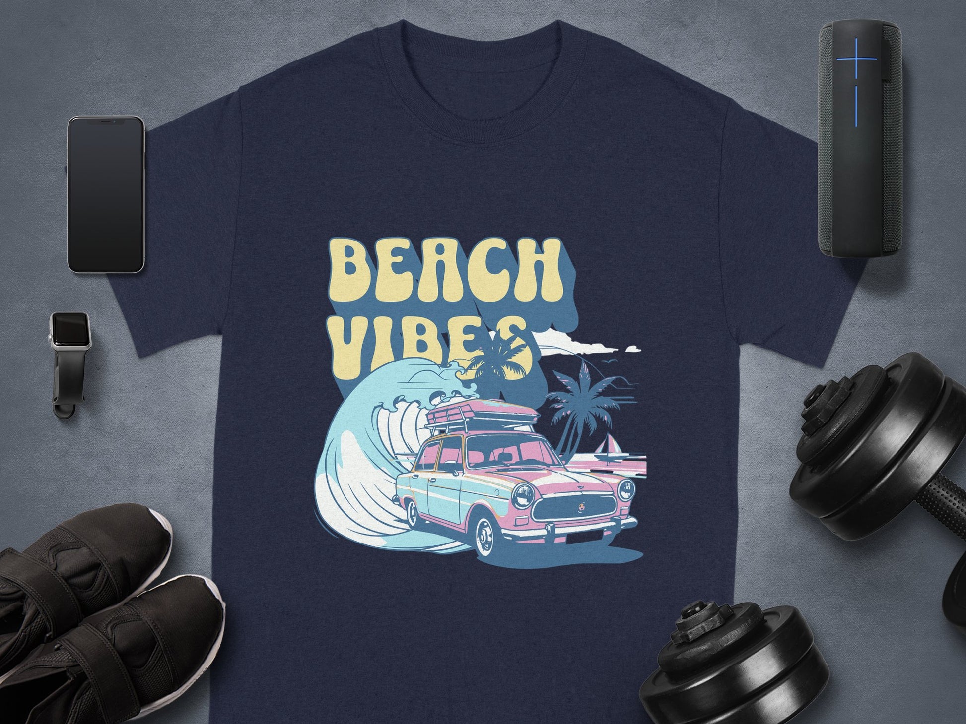 A navy blue Garment Graphics T-shirt shows a pink and blue classic car with surfboards, a wave, palm trees, and Beach Vibes. Nearby are a smartphone, smartwatch, speaker, shoes, and dumbbell on a gray surface.