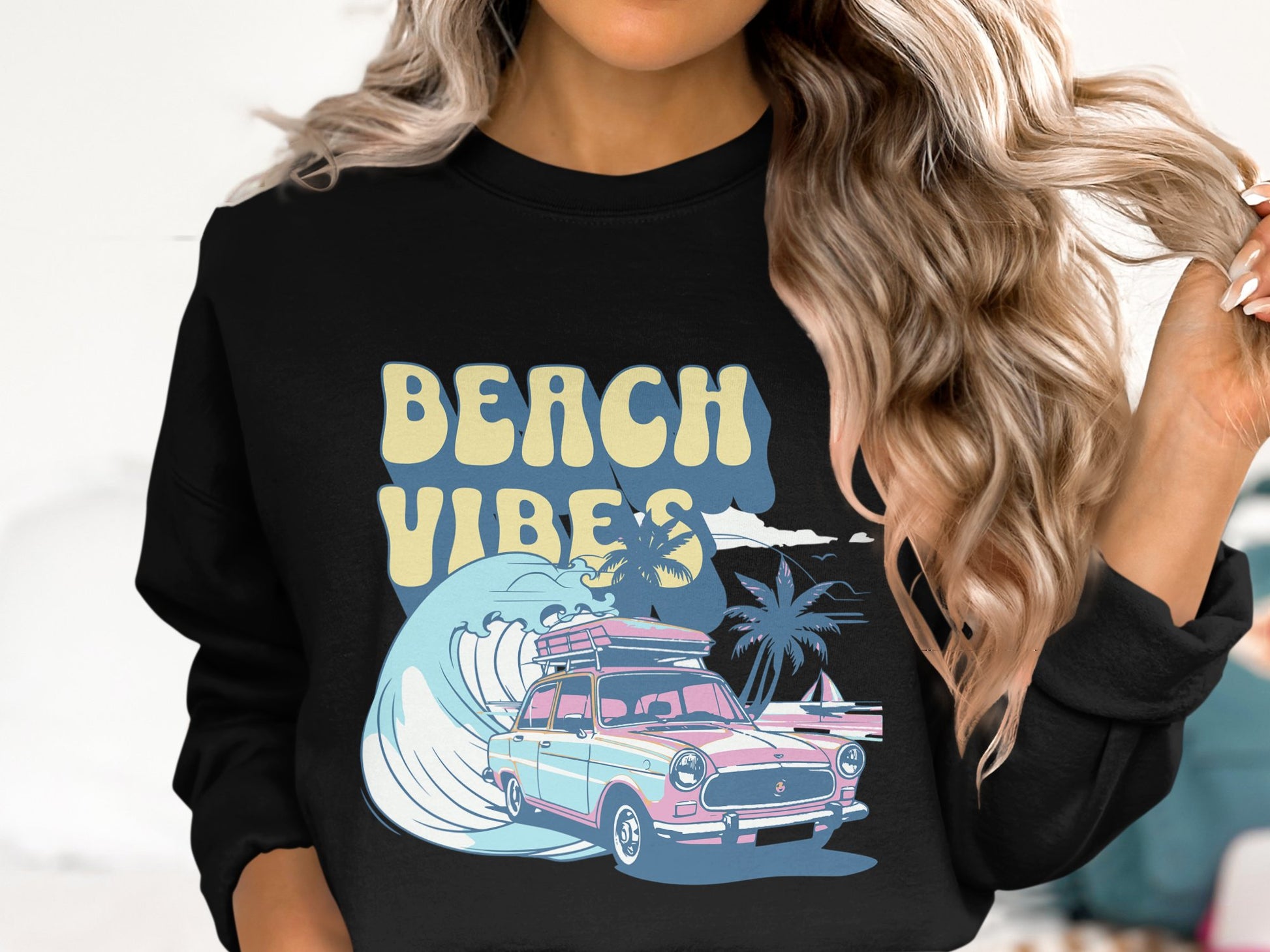 A person with long wavy hair wearing a black Garment Graphics sweatshirt featuring a retro car, surfboard, waves, and palm trees exudes vintage vibes. The BEACH VIBES text captures the fun spirit while the image highlights their upper half.