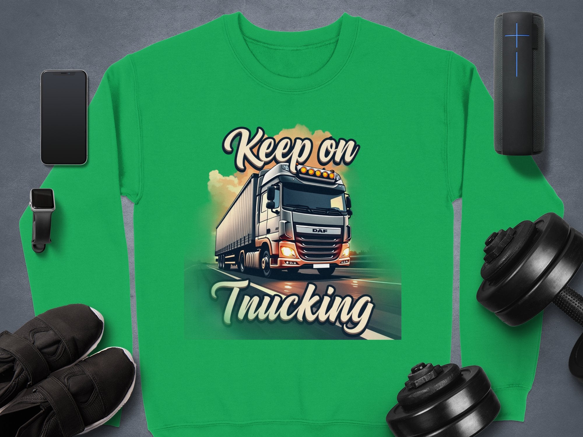 The unisex green graphic sweatshirt by Garment Graphics features a bold truck and Keep on Trucking text, accompanied by essentials like a smartphone, smartwatch, black sneakers, dumbbells, and a portable speaker on a sleek gray surface.