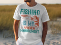 A fishing enthusiast wears a white T-shirt by Garment Graphics with Fishing Is My Anger Management in teal, orange, and red. The design features a fish and hook against a sandy beach backdrop with grasses and a blurry horizon.
