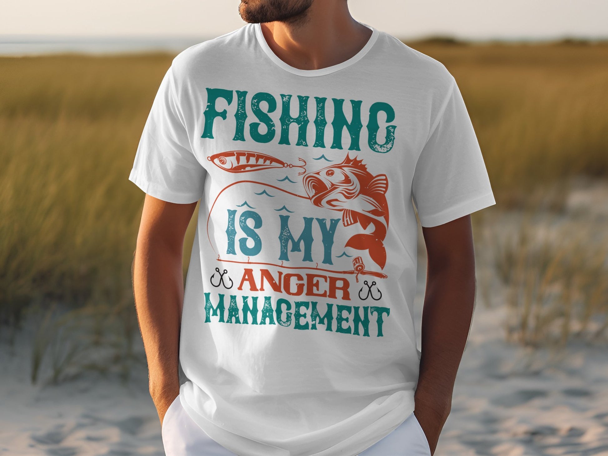 A fishing enthusiast wears a white T-shirt by Garment Graphics with Fishing Is My Anger Management in teal, orange, and red. The design features a fish and hook against a sandy beach backdrop with grasses and a blurry horizon.