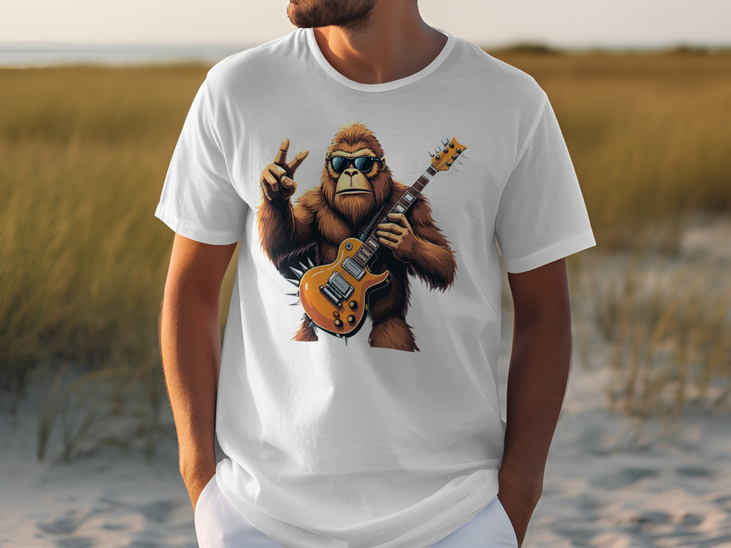 A person is wearing a Garment Graphics white T-shirt showcasing a cartoon gorilla with sunglasses, holding an electric guitar, and making a peace sign against a blurred grassy landscape—ideal for music enthusiasts.