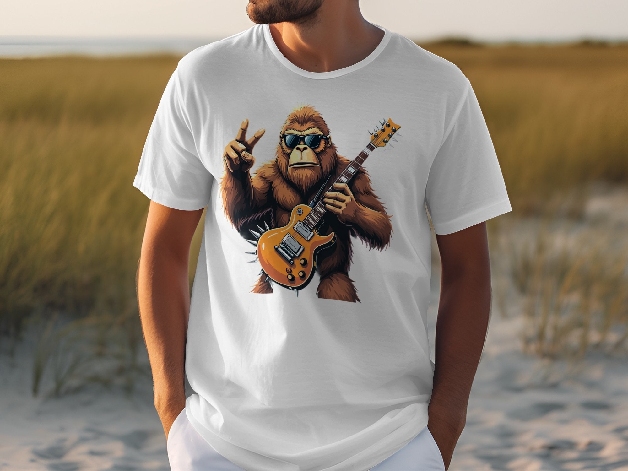 A person is wearing a Garment Graphics white T-shirt showcasing a cartoon gorilla with sunglasses, holding an electric guitar, and making a peace sign against a blurred grassy landscape—ideal for music enthusiasts.