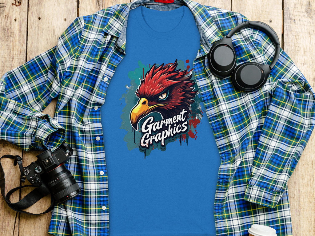 A blue t-shirt with an eagle design and Garment Graphics sits on a plaid shirt on a wooden surface. A camera, headphones, and lidded coffee cup complete the scene, capturing this stylish classic fit ensemble.