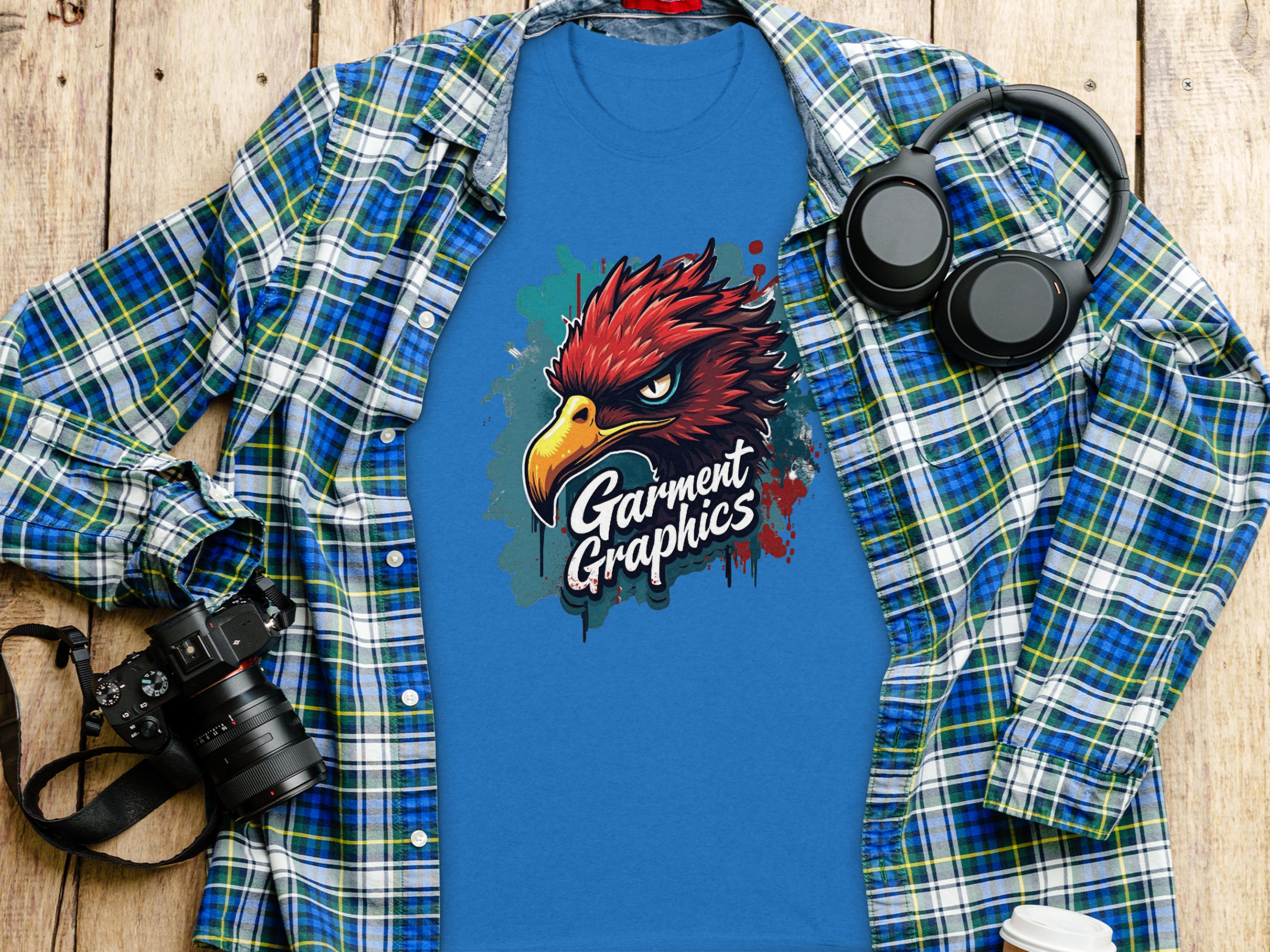 A blue t-shirt with an eagle design and Garment Graphics sits on a plaid shirt on a wooden surface. A camera, headphones, and lidded coffee cup complete the scene, capturing this stylish classic fit ensemble.