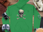 The Garment Graphics green hoodie with a skull and crossbones design stands out on a wooden surface, surrounded by essential accessories: a camera, binoculars, a carabiner, a leather pouch, and sunglasses—ideal for adventure or adding everyday flair.