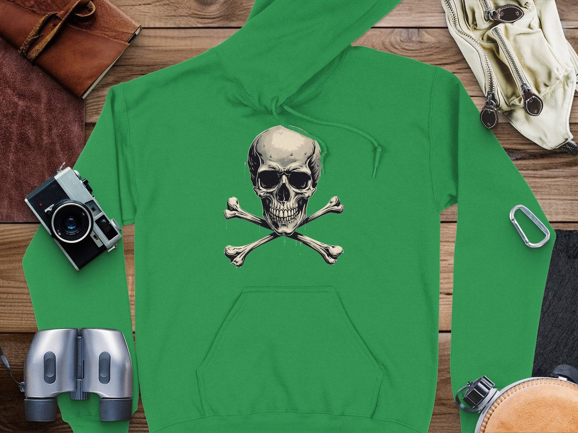 The Garment Graphics green hoodie with a skull and crossbones design stands out on a wooden surface, surrounded by essential accessories: a camera, binoculars, a carabiner, a leather pouch, and sunglasses—ideal for adventure or adding everyday flair.