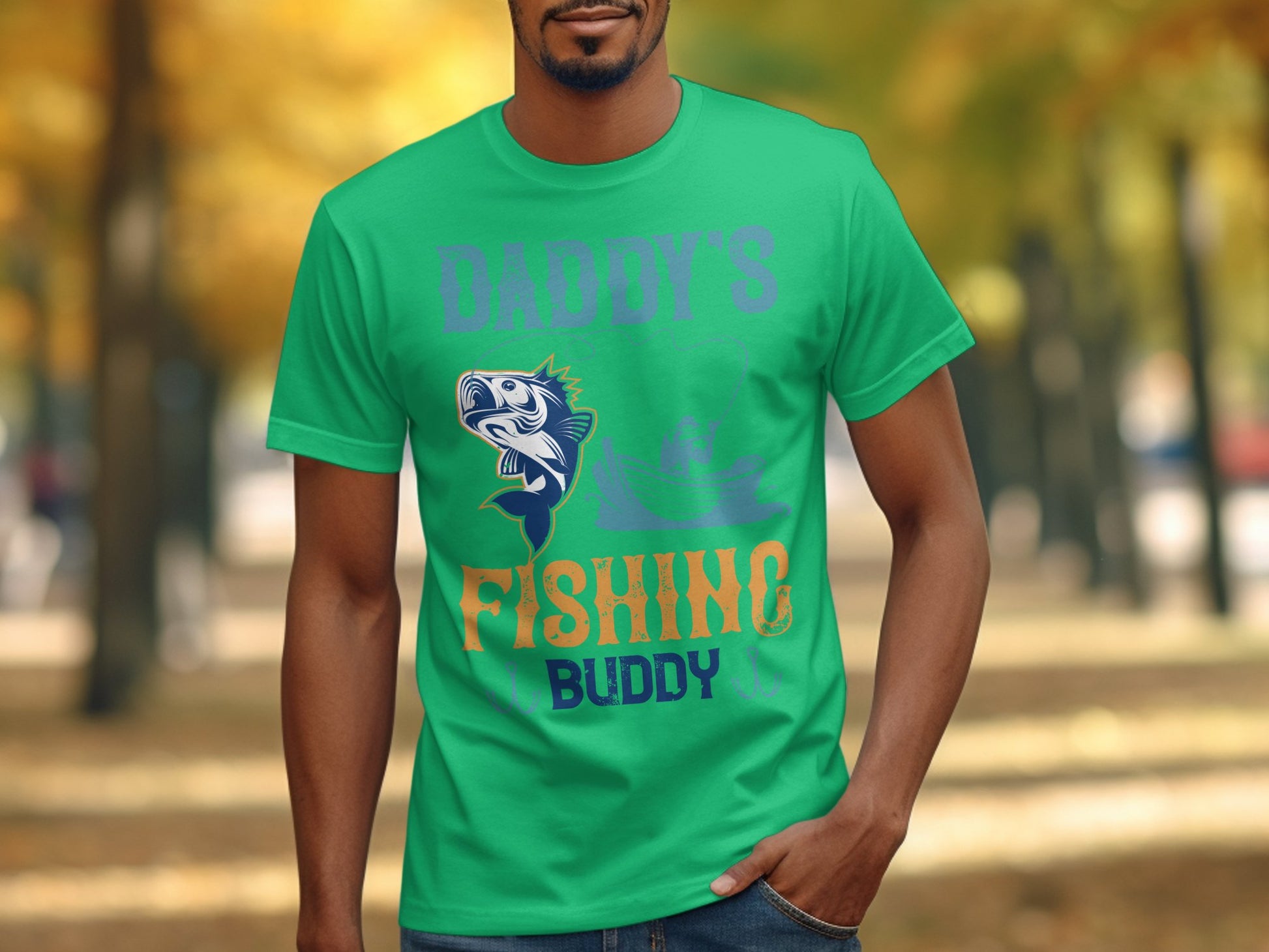 A man wearing a Garment Graphics green graphic print t-shirt with Daddys Fishing Buddy and a whimsical fish design stands outdoors against a backdrop of blurred trees and a winding path, creating an ideal scene for fishing enthusiasts.