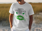 A person wearing a Garment Graphics T-shirt with green lips, clovers, and Happy St. Patricks Day text is outdoors on grass and sand.