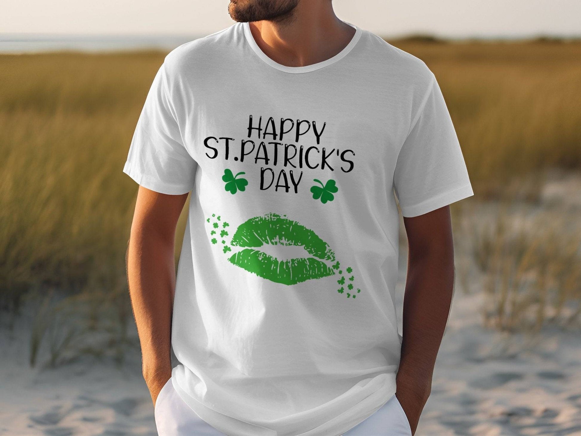 A person wearing a Garment Graphics T-shirt with green lips, clovers, and Happy St. Patricks Day text is outdoors on grass and sand.