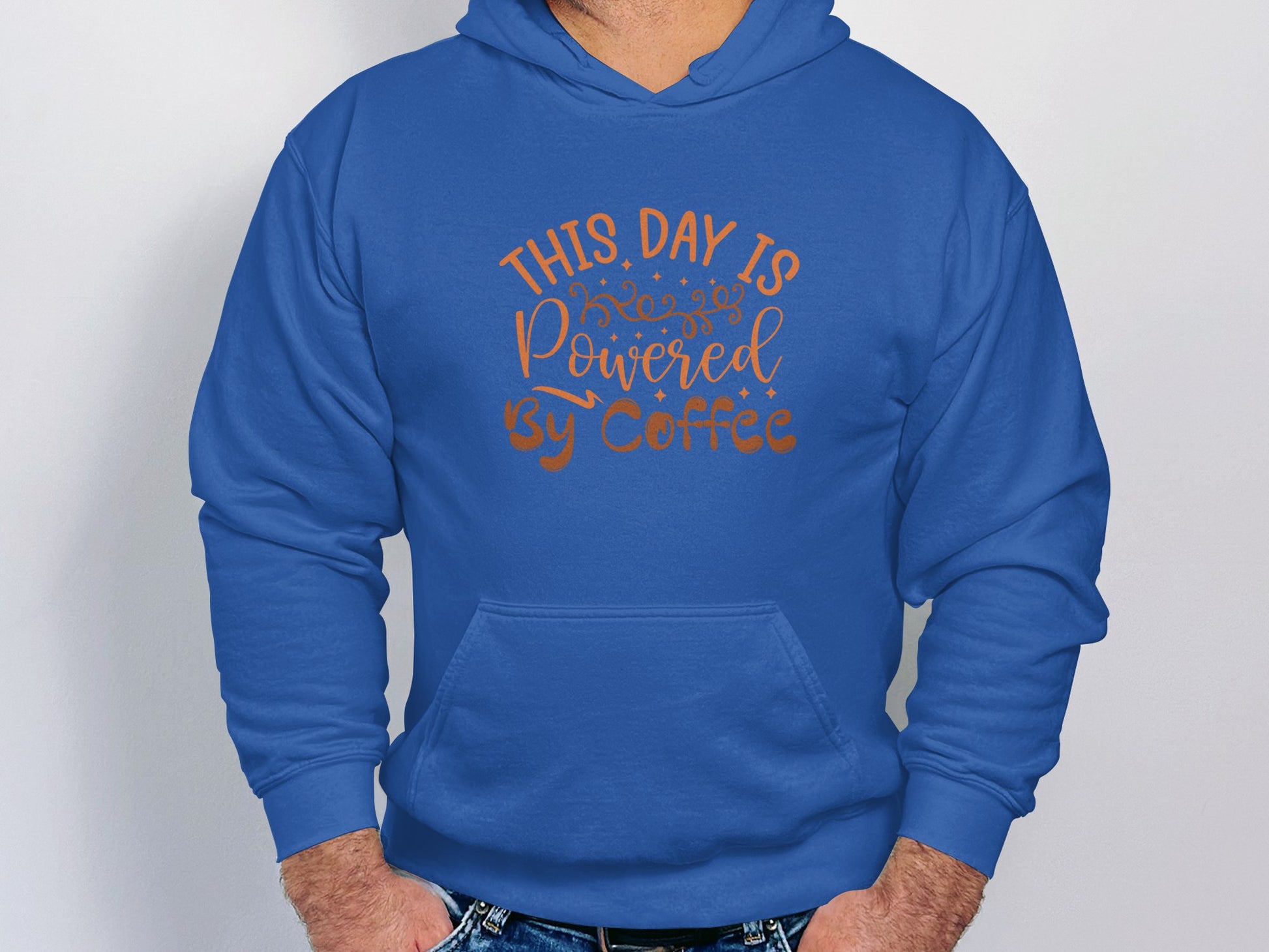 A stylish individual in a Garment Graphics blue hoodie with This day is powered by coffee written in orange, perfect for coffee enthusiasts, stands with hands in the front pocket.