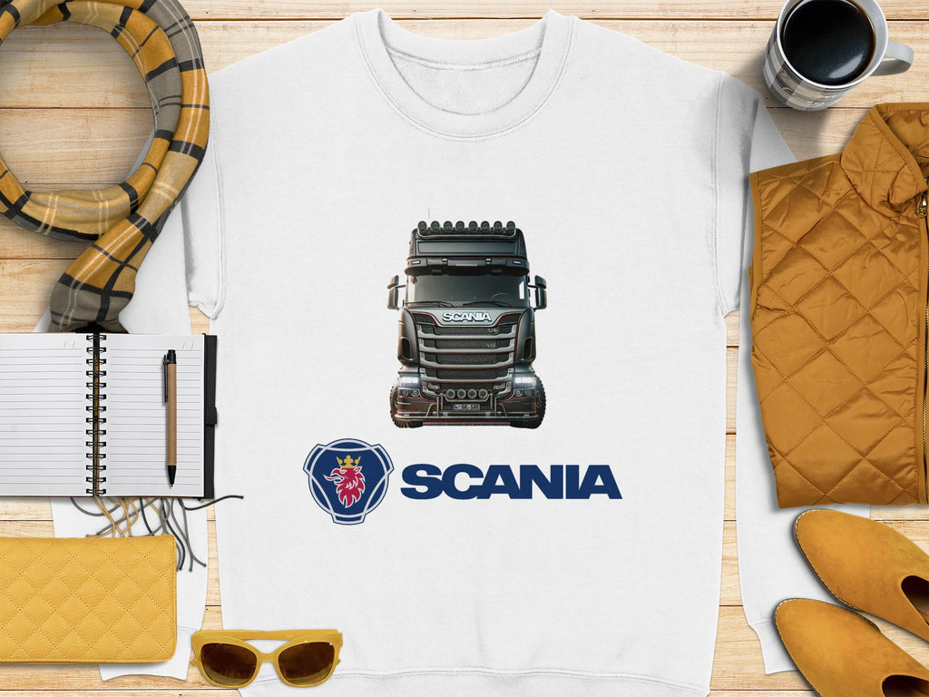 Garment Graphics offers a graphic crewneck sweatshirt with a detailed Scania truck illustration above its iconic logo. This cozy piece is styled on a wooden surface and complemented by accessories like a scarf, coffee cup, quilted jacket, notebook with pen, yellow clutch, and sunglasses. Perfect for truck enthusiasts!.