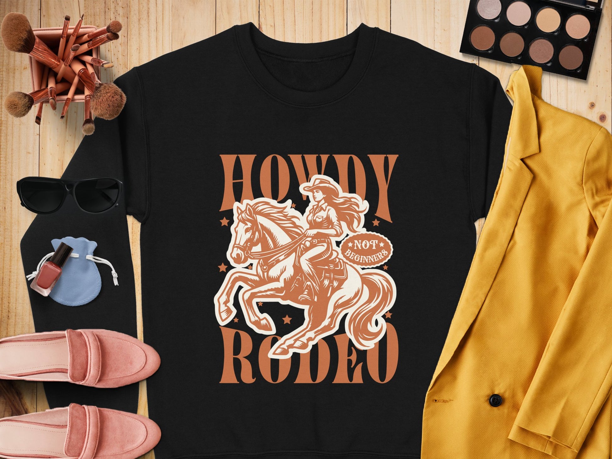 A flat lay features Garment Graphics Howdy Rodeo Sweatshirt with a cowgirl on horseback, styled with rodeo essentials: mustard blazer, pink shoes, sunglasses, makeup brushes, nail polish, blue pouch, and eyeshadow palette on a wooden surface.