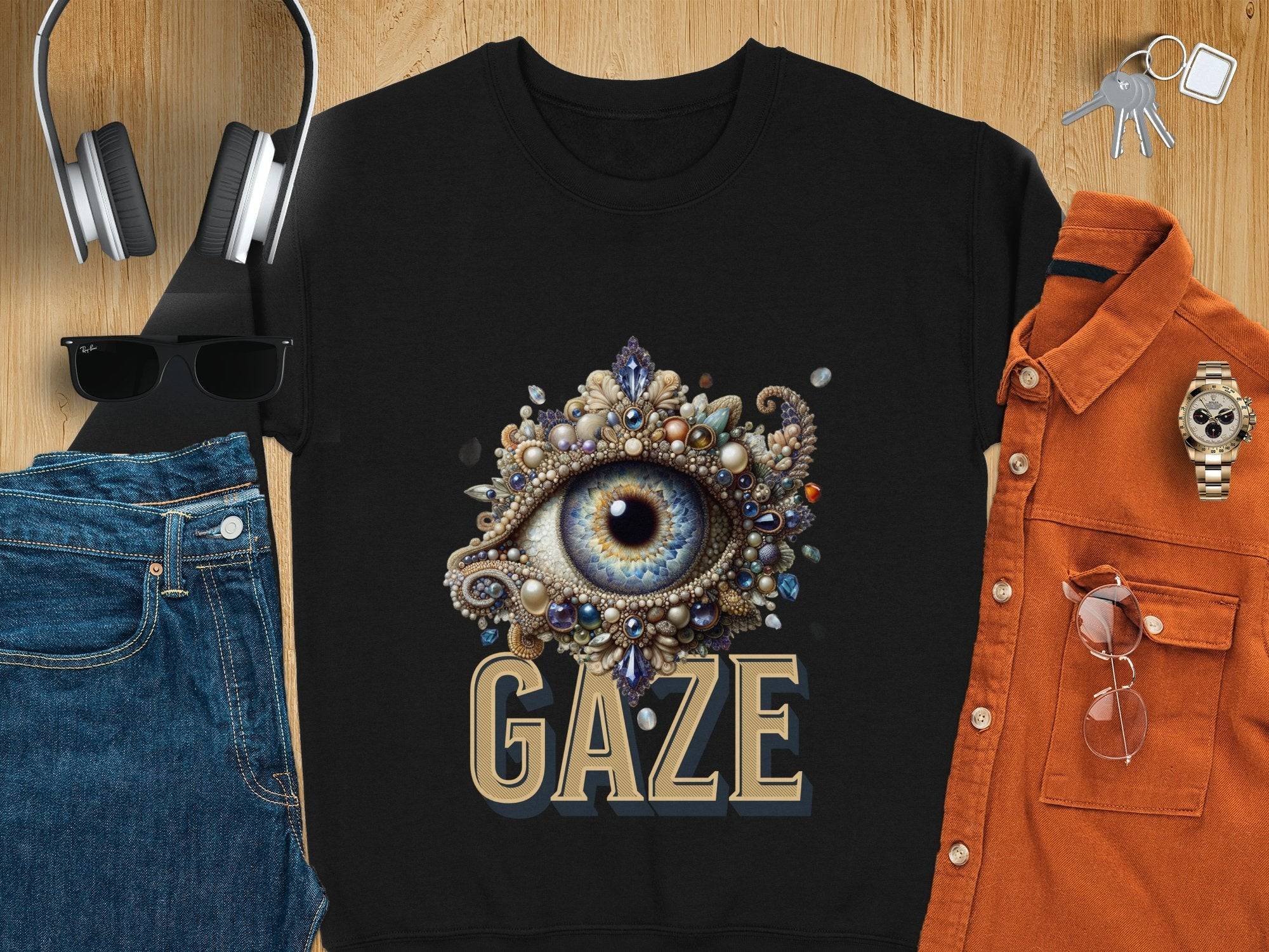 On a wooden surface, a Garment Graphics unisex black t-shirt with an eye design and the word GAZE is styled with jeans, headphones, sunglasses, an orange shirt, a watch, and keys—ideal for any wardrobe.