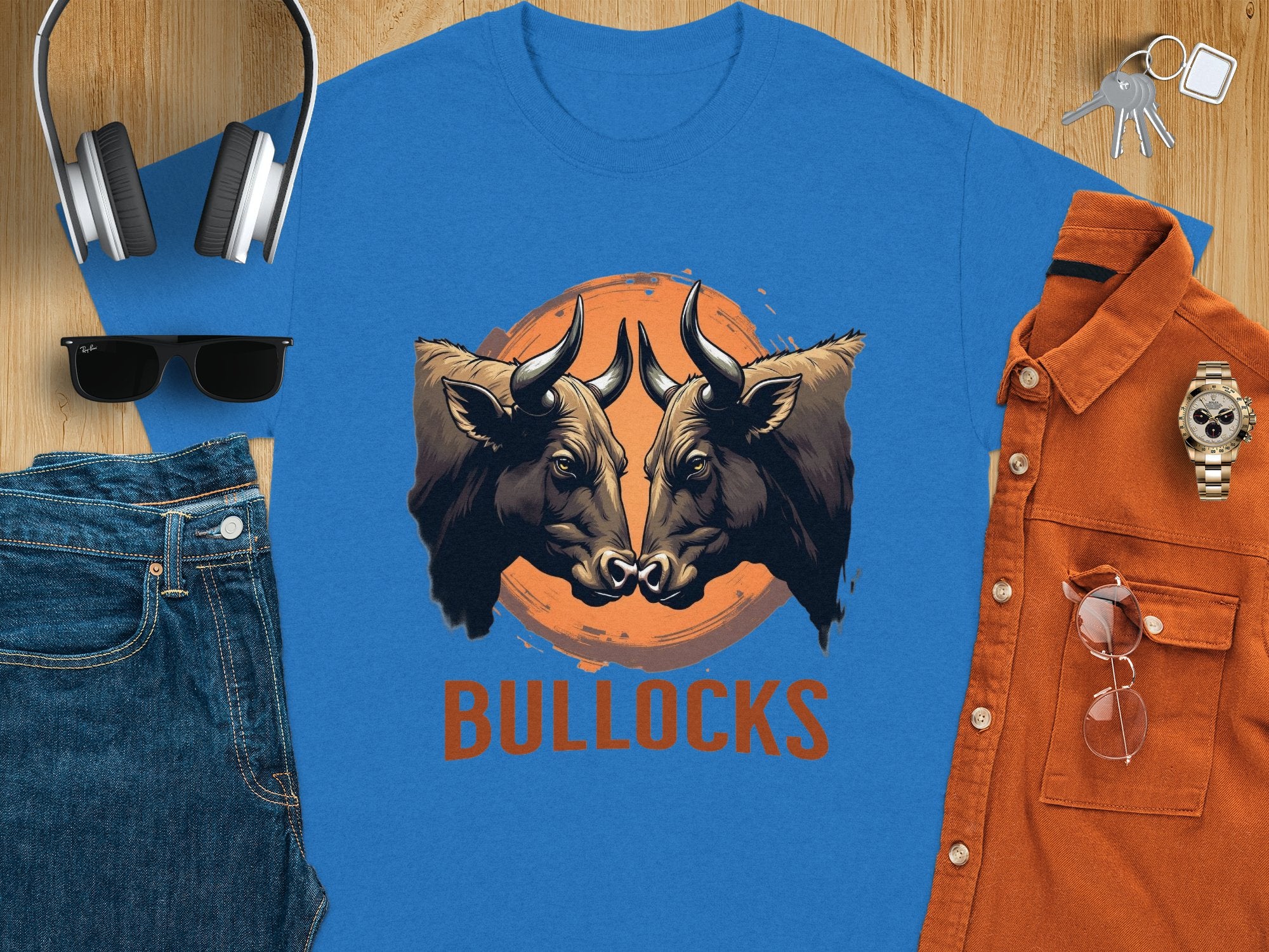 Garment Graphics classic fit blue t-shirt showcases two bull heads facing each other in front of an orange circle with BULLOCKS below. Surrounded by denim jeans, sunglasses, headphones, an orange shirt, keys, and a wristwatch, its perfect for family sizing options.