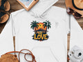 A white Garment Graphics hoodie features a unique style with two palm trees silhouetted against a tropical sunset and the bold text One Love. Its displayed on a wooden surface, surrounded by essentials like a camera, straw hat, notebook, sandals, and sunglasses.