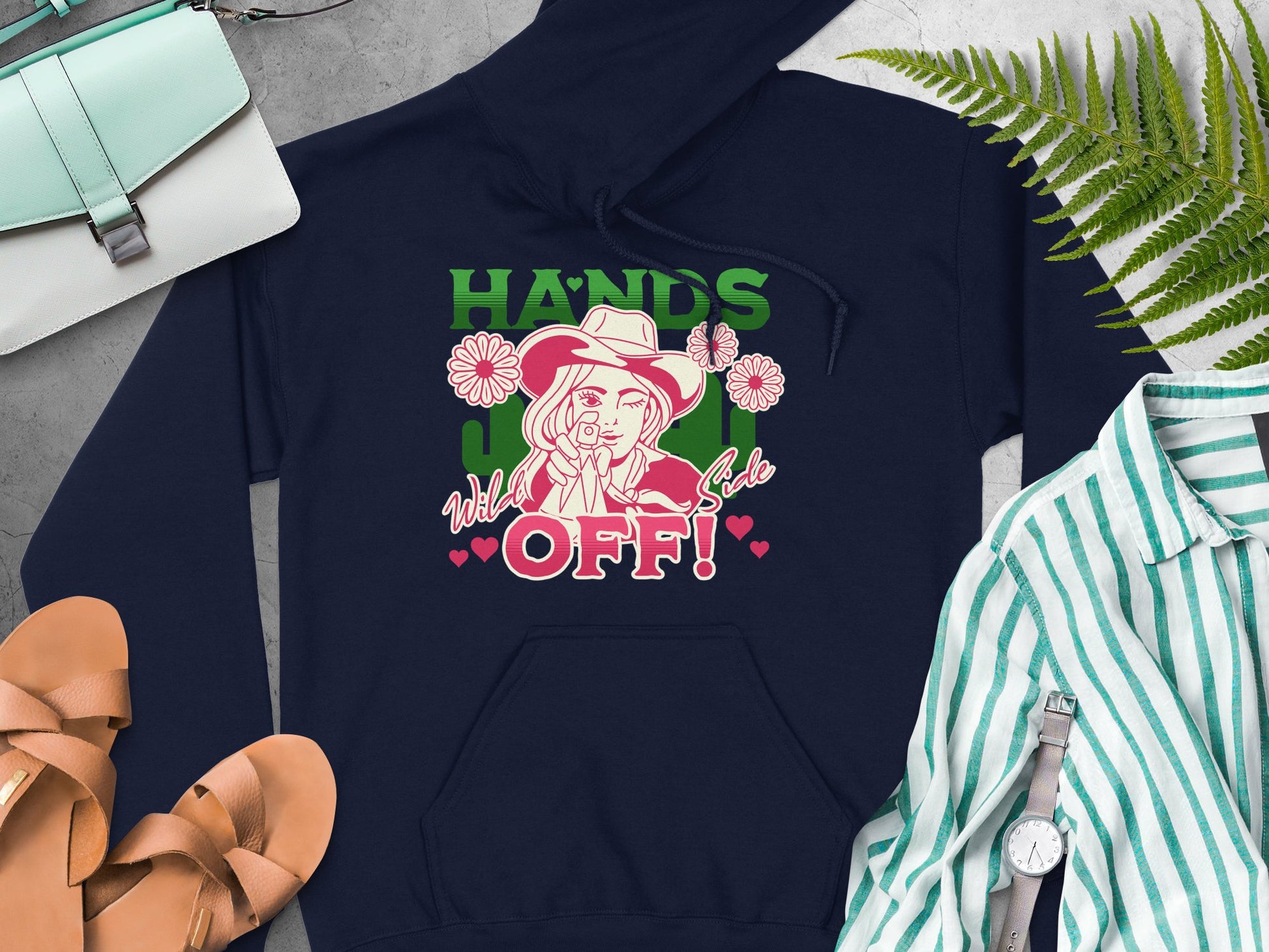 A Garment Graphics navy blue hoodie showcases a vibrant design with a person in a hat and Hands Off! text, surrounded by roses and hearts. Its styled on a surface with a striped shirt, sandals, a watch, and two bags nearby.