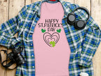 The Garment Graphics pink graphic t-shirt features Happy St. Patricks Day and clover designs overlaid on a blue and green plaid backdrop, surrounded by headphones, a camera, a coffee cup, and a spiral notebook on a wooden surface to create a vibrant festive display.