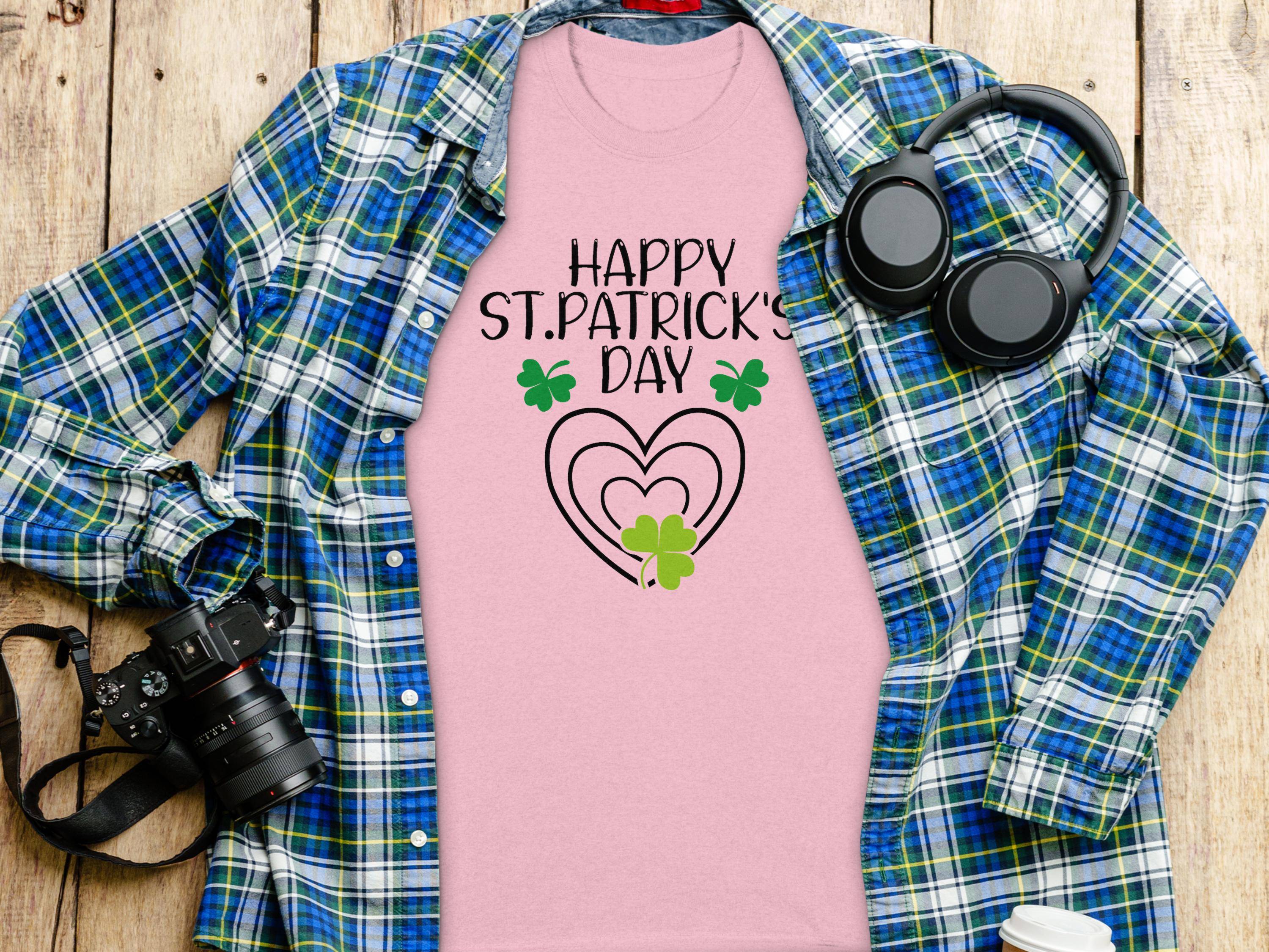 The Garment Graphics pink graphic t-shirt features Happy St. Patricks Day and clover designs overlaid on a blue and green plaid backdrop, surrounded by headphones, a camera, a coffee cup, and a spiral notebook on a wooden surface to create a vibrant festive display.