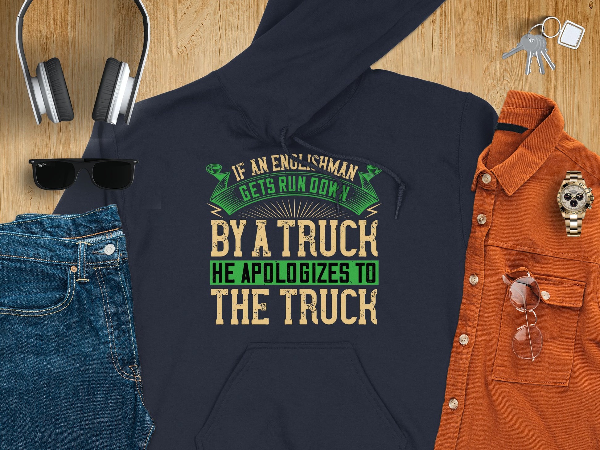 A flat lay of a Garment Graphics navy hoodie with the witty quote, If an Englishman gets run down by a truck, he apologizes to the truck, surrounded by jeans, sunglasses, headphones, a brown shirt, and a watch on wood—showcasing English manners humor.