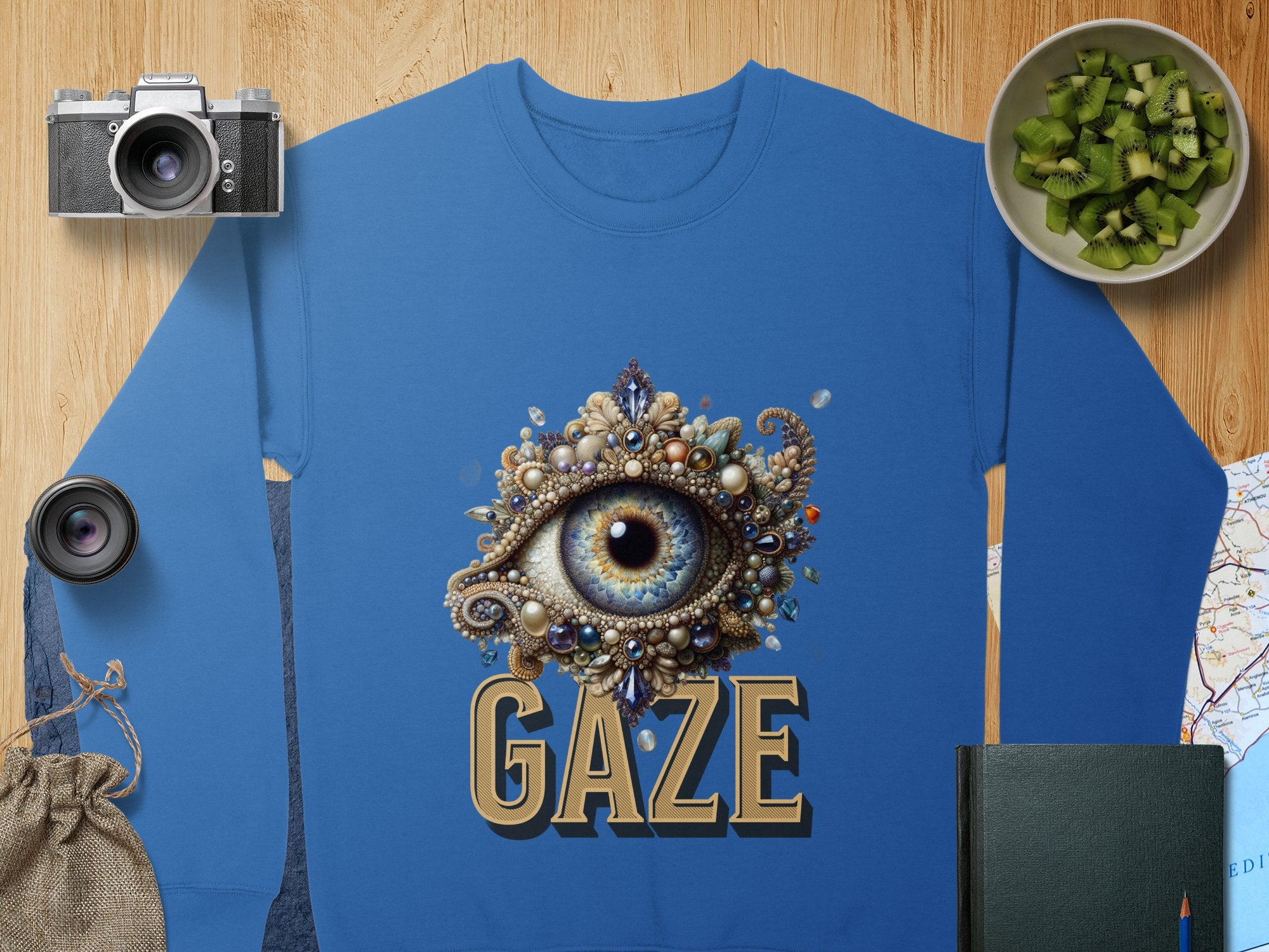A unisex blue graphic sweatshirt by Garment Graphics features an ornate jeweled eye design with the word GAZE beneath. Displayed on wood, it is surrounded by a camera, lens, chopped kiwi, pouch, book, and map. .