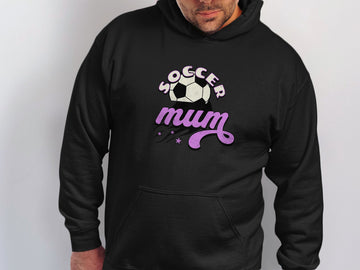 A person stands wearing a cozy black hoodie from Garment Graphics, featuring Soccer Mum in white and purple with a soccer ball graphic and star accents. This classic fit hoodie is perfect for those who embrace their sporty spirit with style.