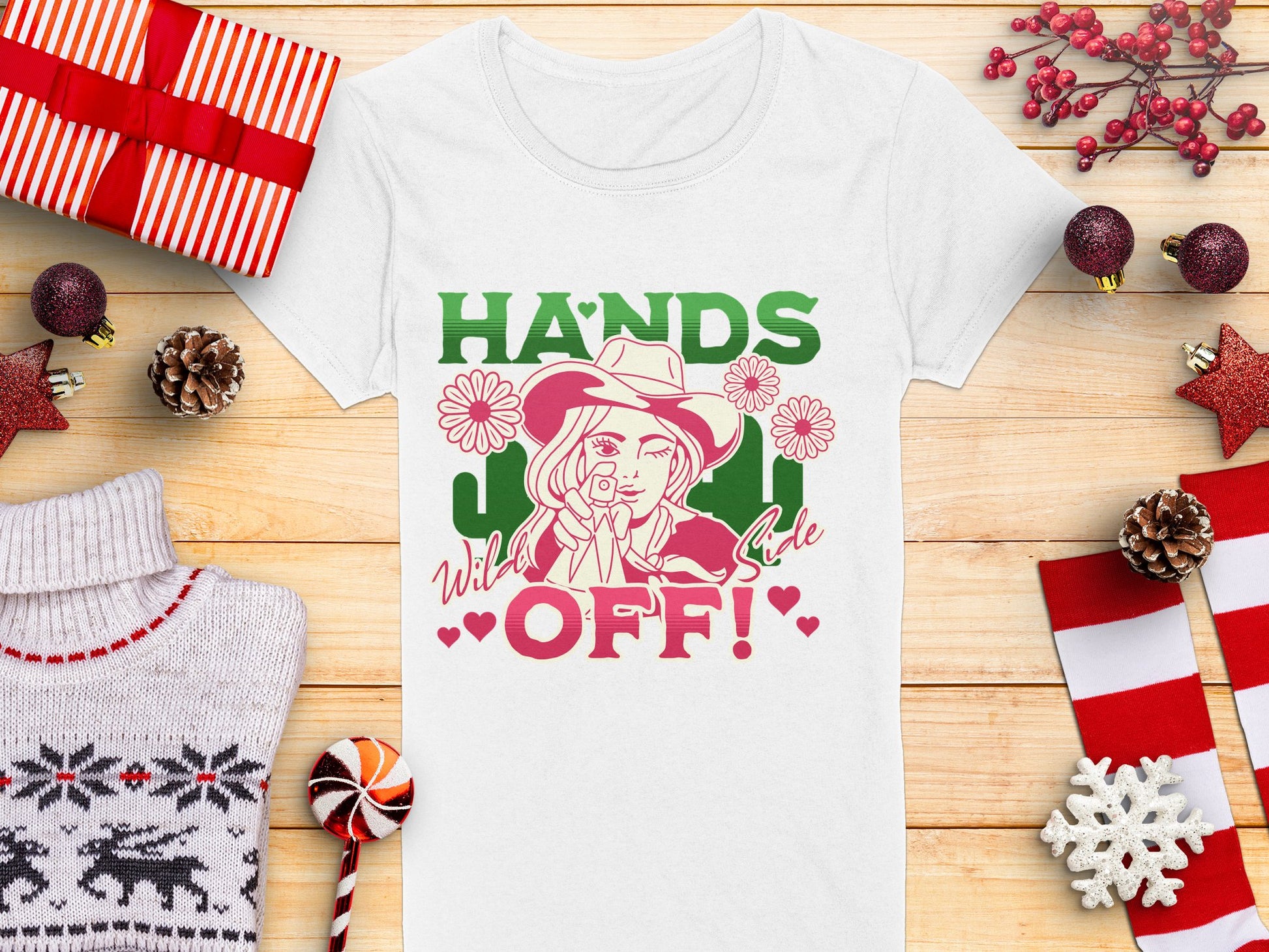 This bold Garment Graphics cowboy T-shirt showcases a person in a cowboy hat surrounded by daisies with Hands Off! in vibrant green and red. Set on wood, its framed by Christmas decorations, a cozy sweater, and wrapped gifts for a unique holiday style.