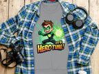 A grey T-shirt from Garment Graphics, showcasing a cartoon superhero and Hero Time! text, rests on wood. Its surrounded by a blue-green plaid shirt, headphones, a camera, and a white cup, all enhancing its versatile unisex appeal.