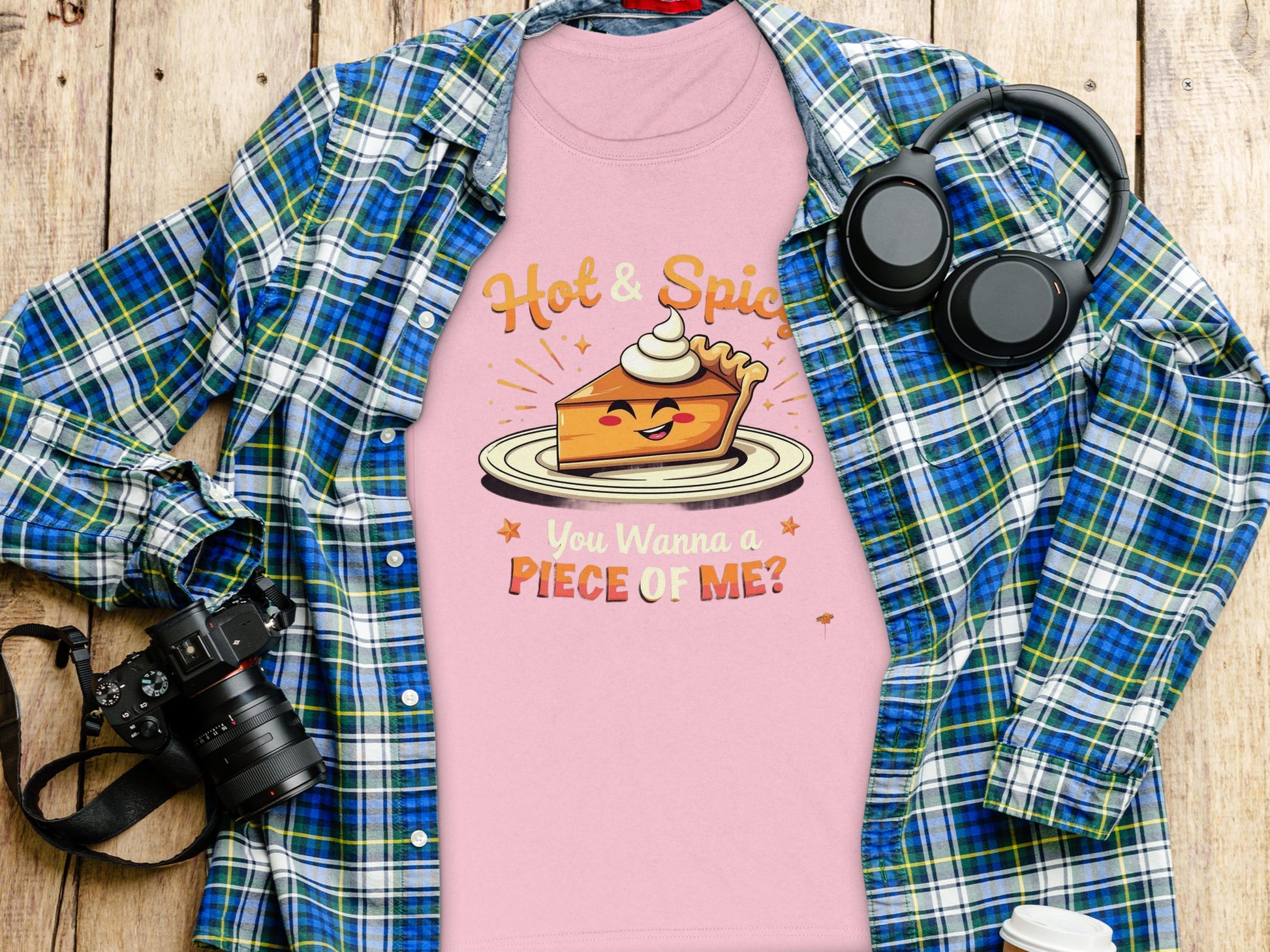 A Garment Graphics plaid shirt, a pink T-shirt with Hot & Spice, You Wanna a Piece of Me? graphic, headphones, and a camera lie on wood. Perfectly sized for women and ideal for casual outings.