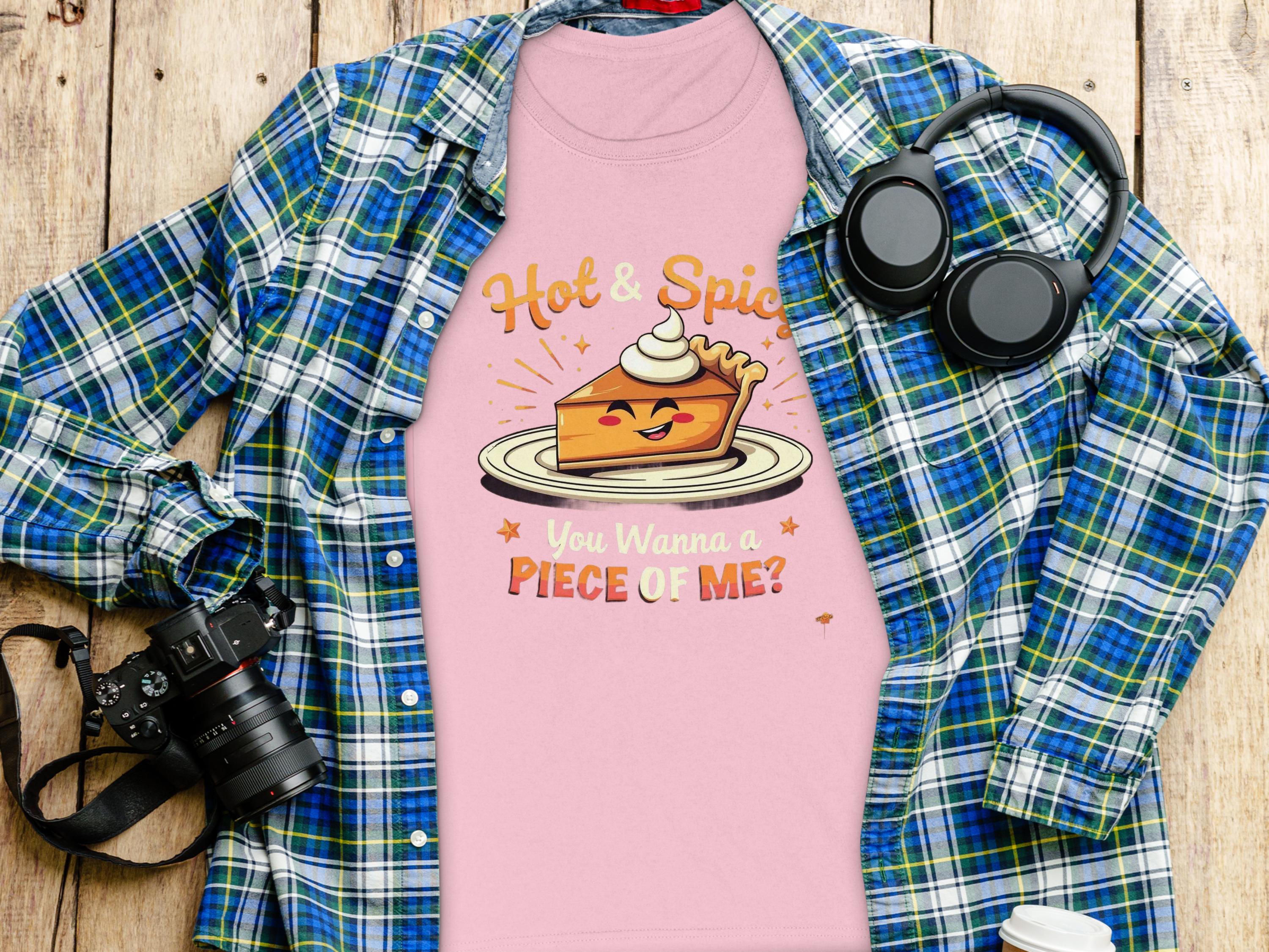 A Garment Graphics plaid shirt, a pink T-shirt with Hot & Spice, You Wanna a Piece of Me? graphic, headphones, and a camera lie on wood. Perfectly sized for women and ideal for casual outings.