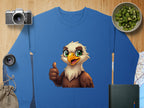 This blue sweatshirt from Garment Graphics features a cheerful cartoon eagle giving a thumbs-up. It is unisex sized and displayed on a wooden surface, surrounded by a camera, lens, sliced kiwi bowl, jute bag, and an intriguing book with a map.