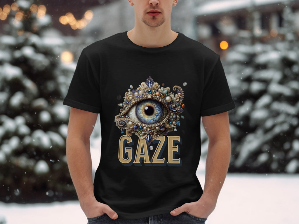 In a snowy outdoor scene, someone wears a classic fit black graphic T-shirt from Garment Graphics. It features an ornate eye design with colorful jewels and GAZE printed below. Snow-covered trees and buildings are blurred in the background.