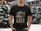 In a snowy outdoor scene, someone wears a classic fit black graphic T-shirt from Garment Graphics. It features an ornate eye design with colorful jewels and GAZE printed below. Snow-covered trees and buildings are blurred in the background.