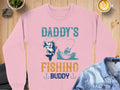 A cozy scene features a Garment Graphics pink graphic sweatshirt on wood, labeled Daddys Fishing Buddy with fishing imagery. A latte, rocks, and a denim jacket add charm for outdoor lovers.