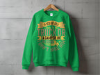 A green sweatshirt by Garment Graphics hangs on the wall, declaring: If hes got golf clubs in his truck or a camper in his driveway, I dont hire him. It features gold and tan lettering with a small image of a truck towing a camper—ideal for outdoor enthusiasts.