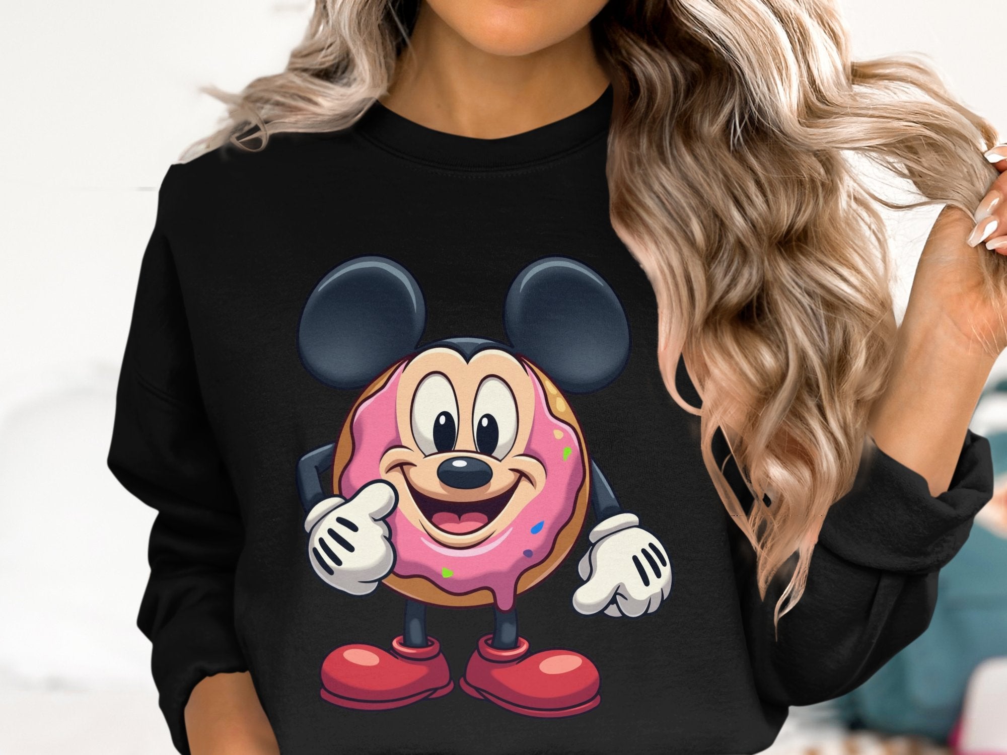 The individual in a black sweatshirt from Garment Graphics showcases a playful donut character with mouse ears, a pink sprinkle face, and red shoes. Highlighting whimsical charm by clasping their wavy hair, this unisex piece ensures everyone can enjoy the fun design.