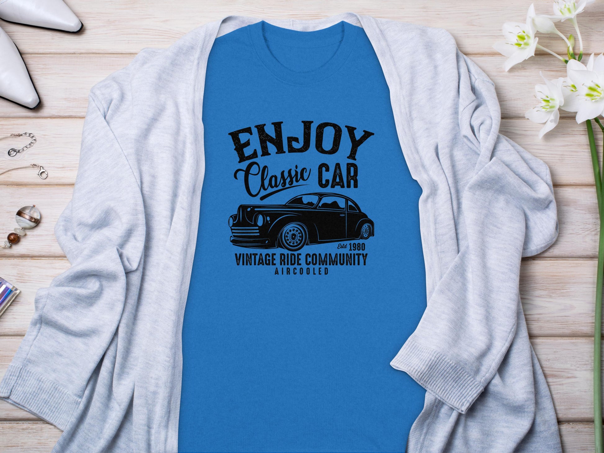 The Garment Graphics blue T-shirt features the text Enjoy Classic Car, Vintage Ride Community, Aircooled with a car illustration. Its ideal for car enthusiasts and is styled on a wooden surface with a light gray cardigan, white high heels, jewelry, and flowers.