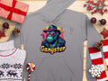 This gray hoodie by Garment Graphics features a gangster ape in sunglasses, a hat, and smoking above the word Gangster. Ideal for hipster urban style fans, it is photographed with wrapped gifts, candy, pinecones, and decorative stars on a wooden surface.